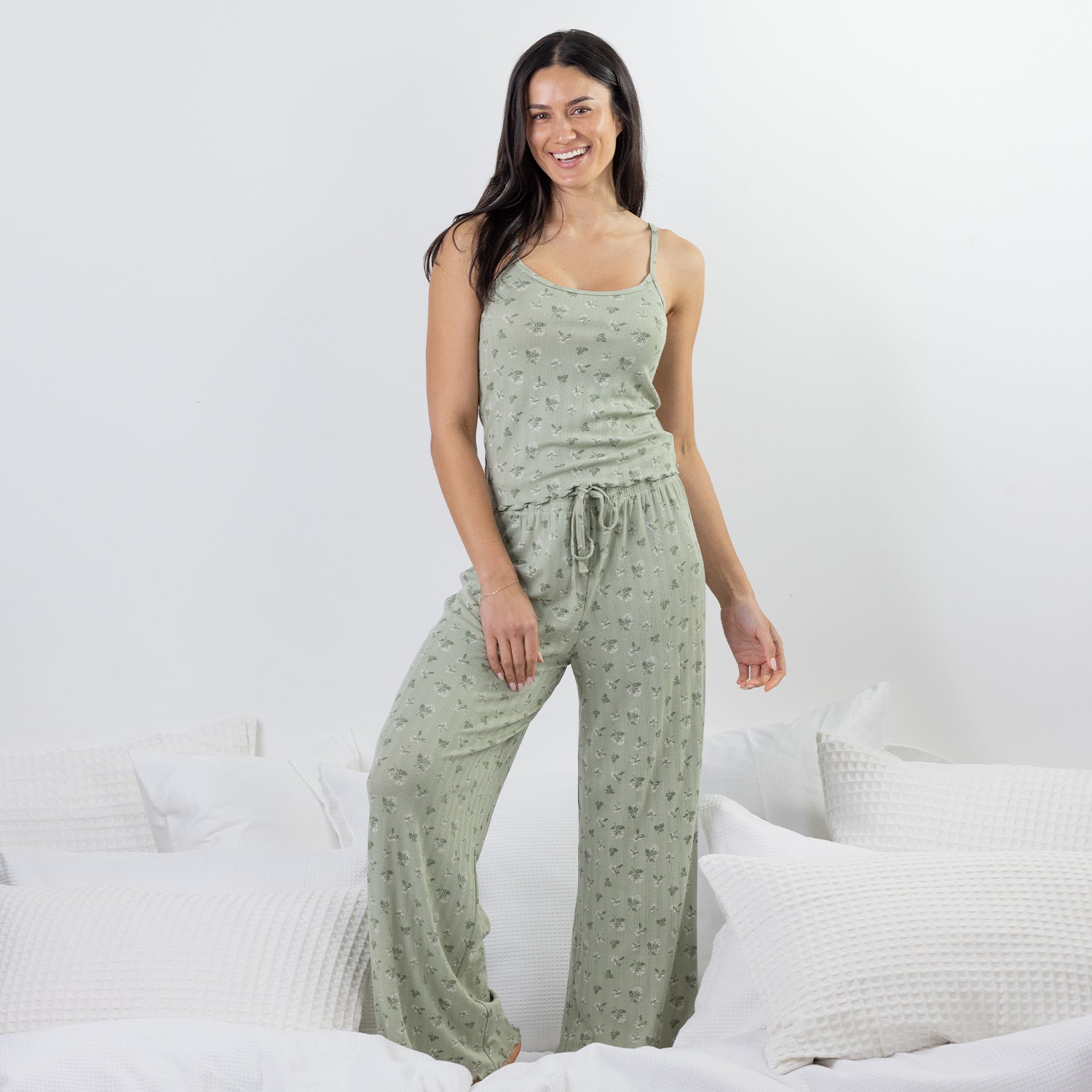 A woman modeling a sage green two-piece pajama set with a small floral print, consisting of a camisole top and matching wide-leg pants, posed on a bed with white pillows.