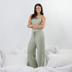 A woman stands on a bed wearing a light sage green pajama set with a small floral print. The set includes a spaghetti-strap tank top and matching wide-leg pj pants with a drawstring waist.