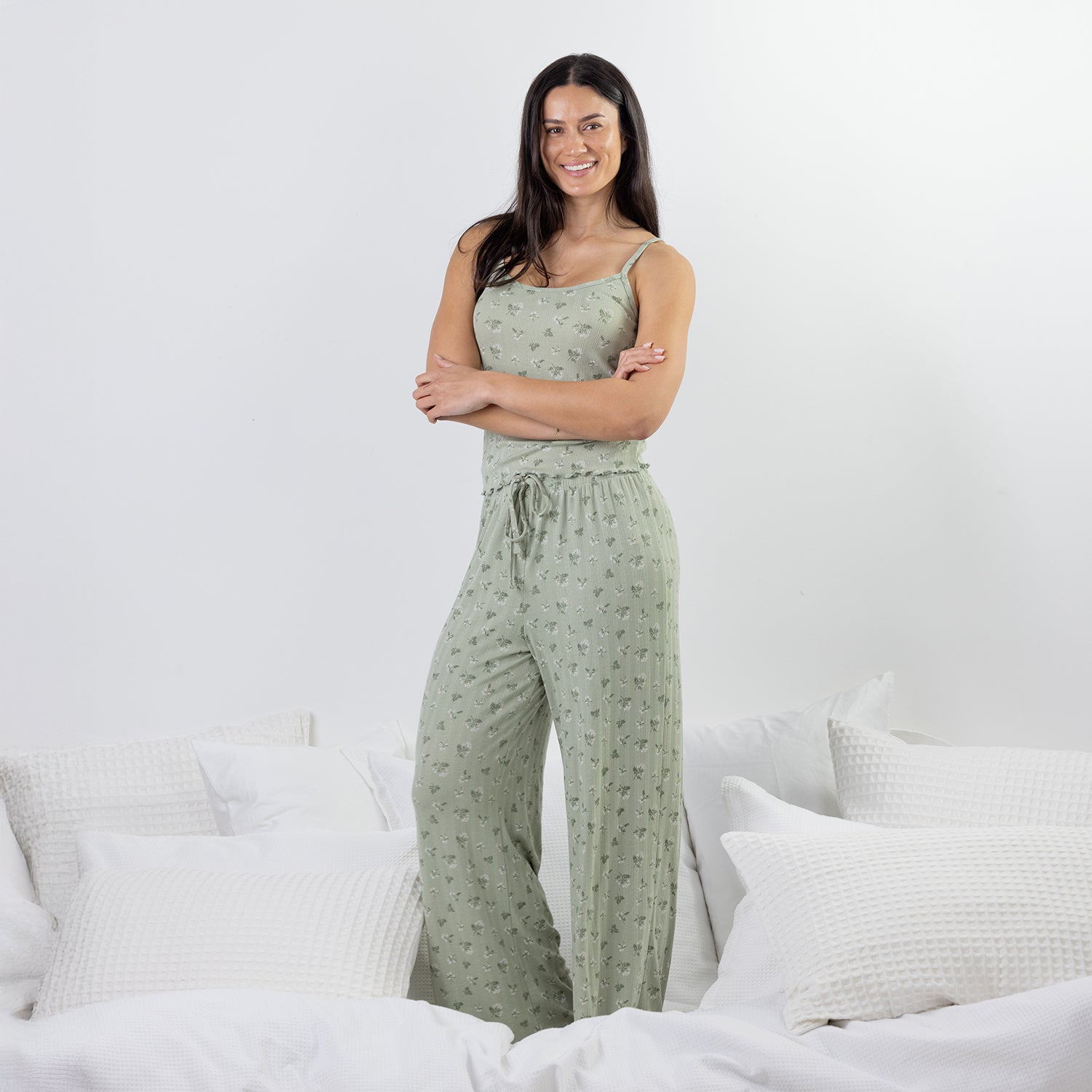 A woman stands on a bed wearing a light sage green pajama set with a small floral print. The set includes a spaghetti-strap tank top and matching wide-leg pj pants with a drawstring waist.