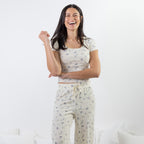 A woman modeling a matching two-piece pajama set consisting of a short-sleeve crop top and drawstring pants, both in a cream color with a small blue floral print, set against a bright white background.