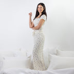 A woman stands on a bed wearing a cream-colored pj setwith a small blue floral pattern. The room is decorated with white bedding and pillows against a white wall.