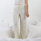 A person wearing cream-colored drawstring pj pants with a small blue floral print, standing on a bed with white bedding and pillows.