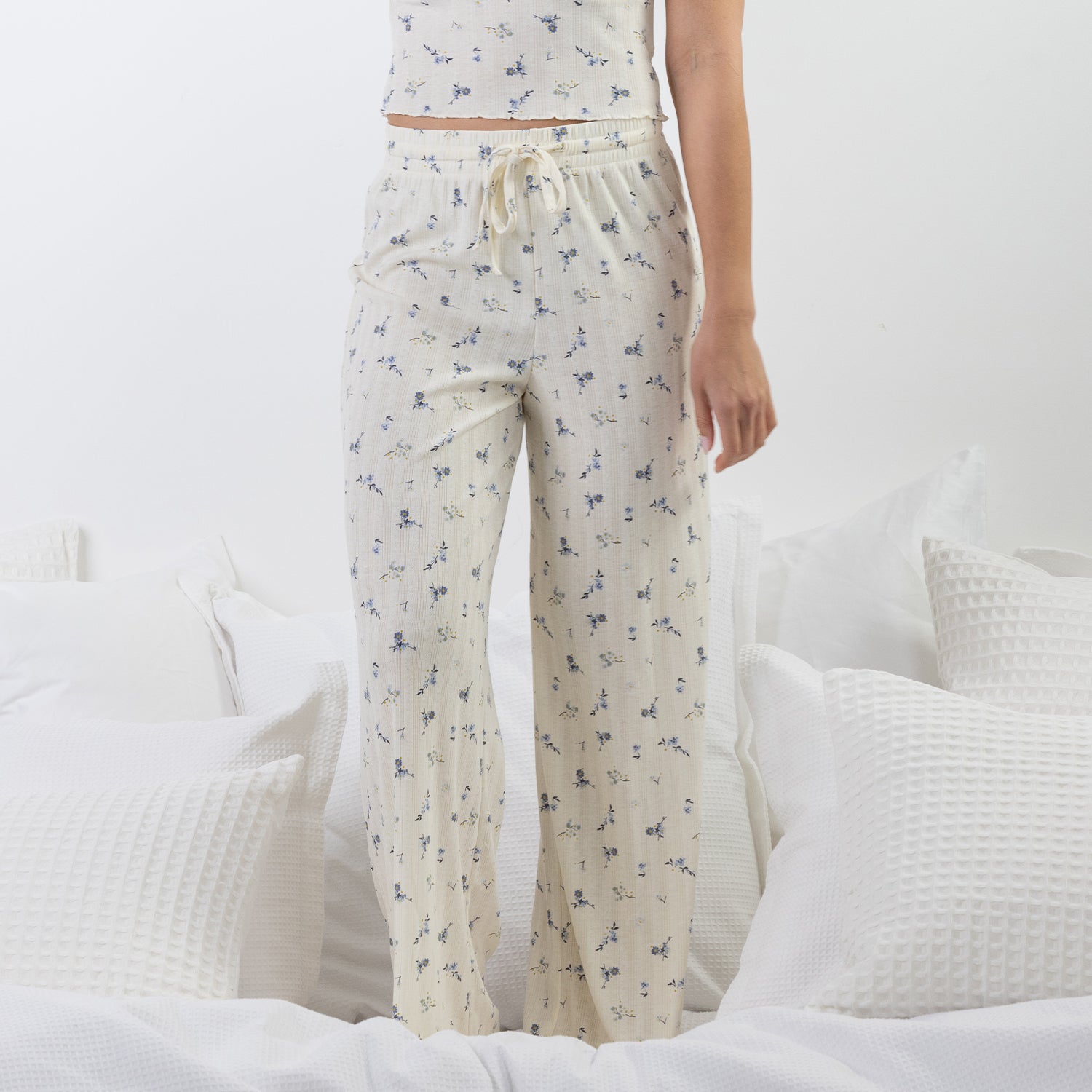 A person wearing cream-colored drawstring pj pants with a small blue floral print, standing on a bed with white bedding and pillows.