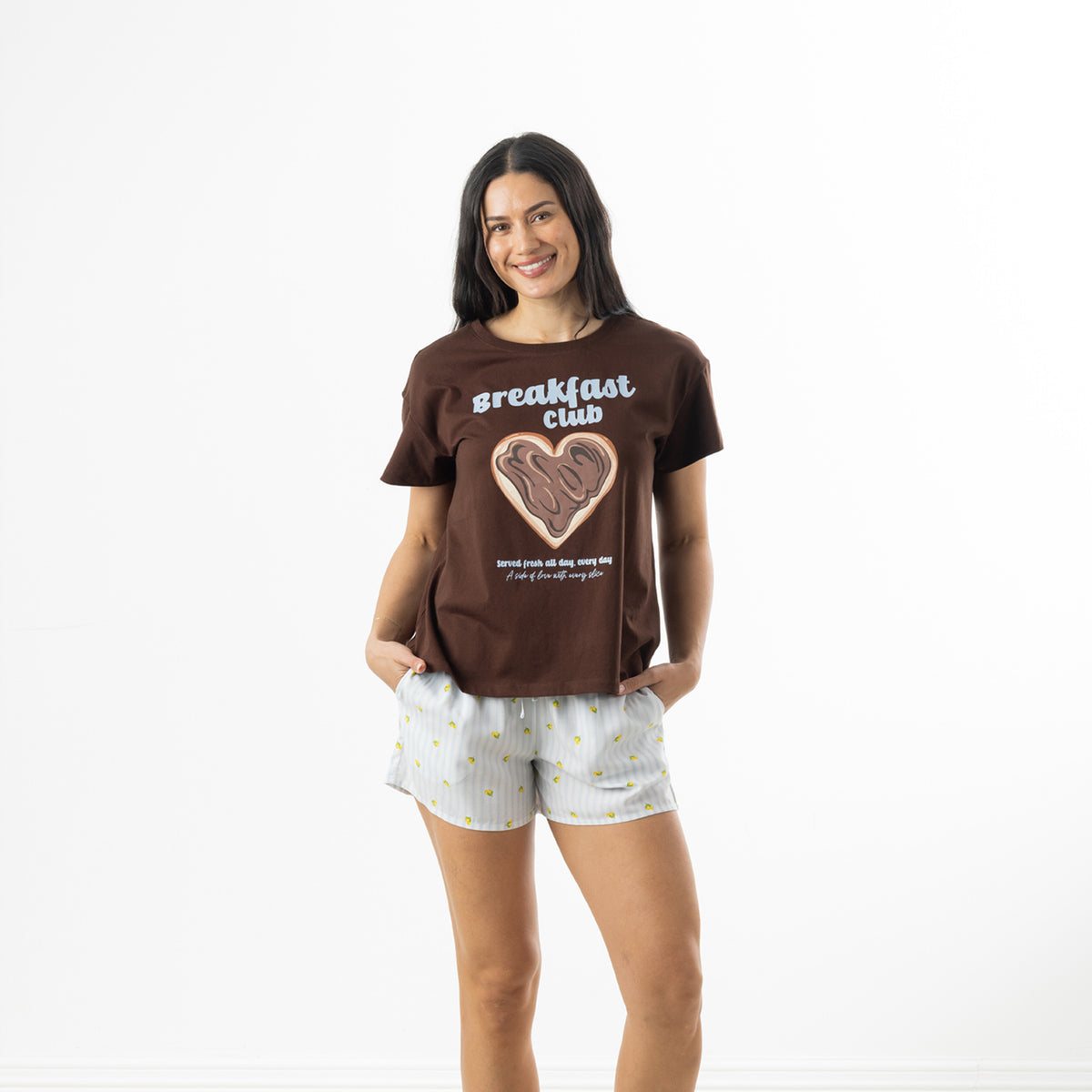 Model wearing a chocolate brown bb&b Sleep Heart Toast Print Sleep Tee with a breakfast club graphic, paired with light blue striped pyjama shorts.