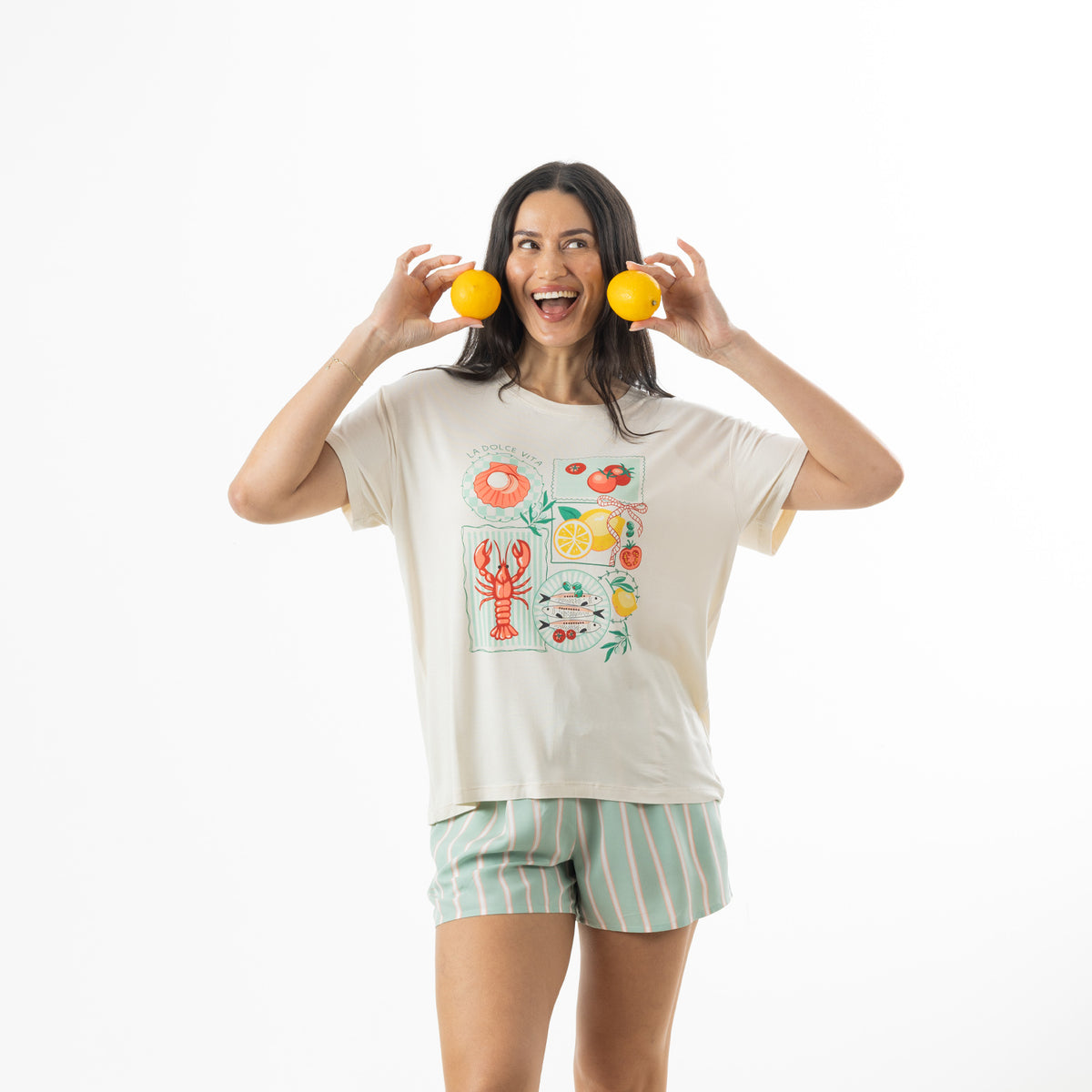 Model wearing an off-white bb&b Sleep Lobster Print Sleep Tee with a colourful Mediterranean-inspired graphic, paired with mint green striped pyjama shorts while holding two lemons.