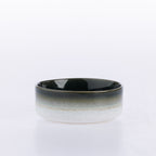 A wide ceramic bowl with a glossy black interior and a speckled white exterior featuring a subtle gold band around the middle, set against a plain white background.