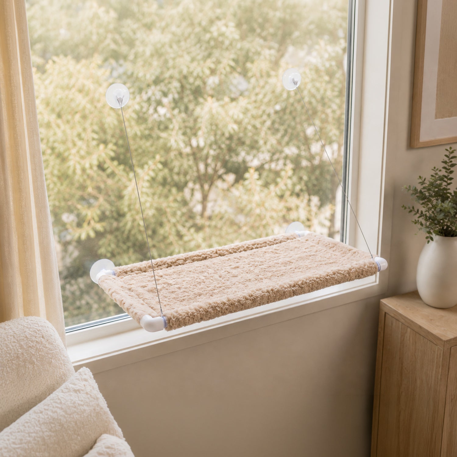 A beige carpeted window perch with white suction cup mounts attached to a window in a bright room, with a wooden side table and potted plant nearby.