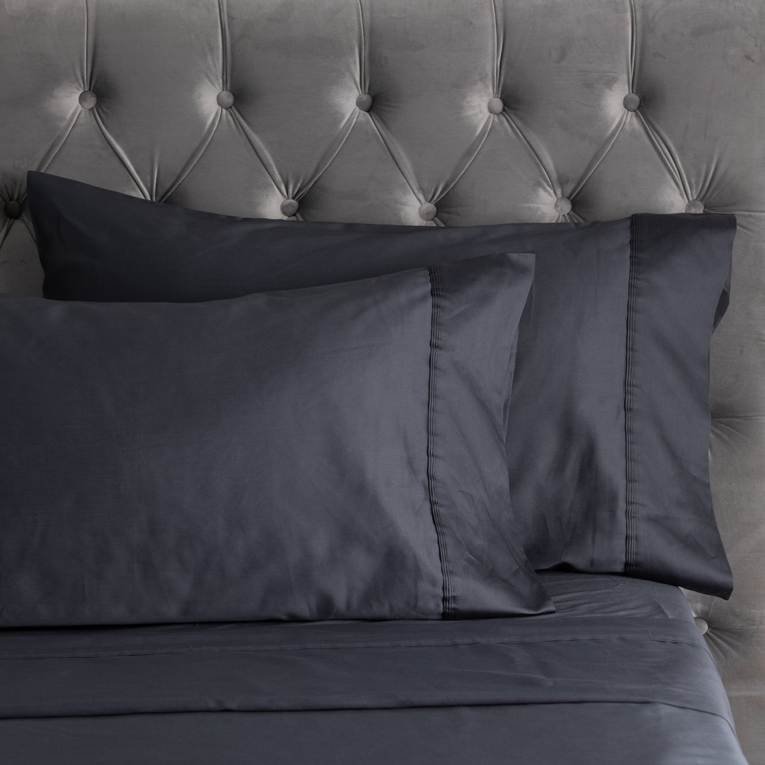A close-up of smooth graphite sheets and matching pillowcases with elegant stitched cuff detailing, styled against a plush tufted velvet headboard.