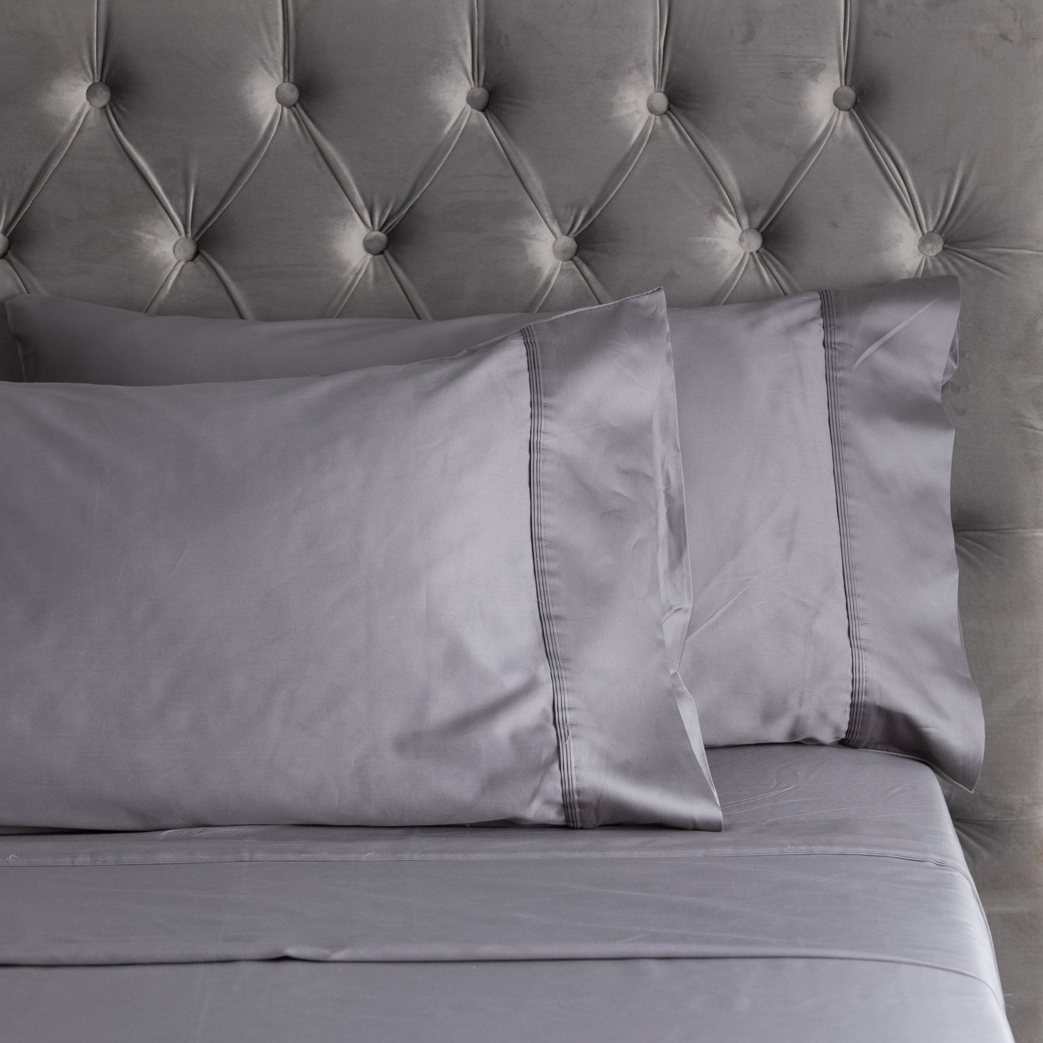 A close-up of smooth grey sheets and matching pillowcases with elegant stitched cuff detailing, styled against a plush tufted velvet headboard.