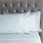 A close-up of smooth ice blue sheets and matching pillowcases with elegant stitched cuff detailing, styled against a plush tufted velvet headboard.