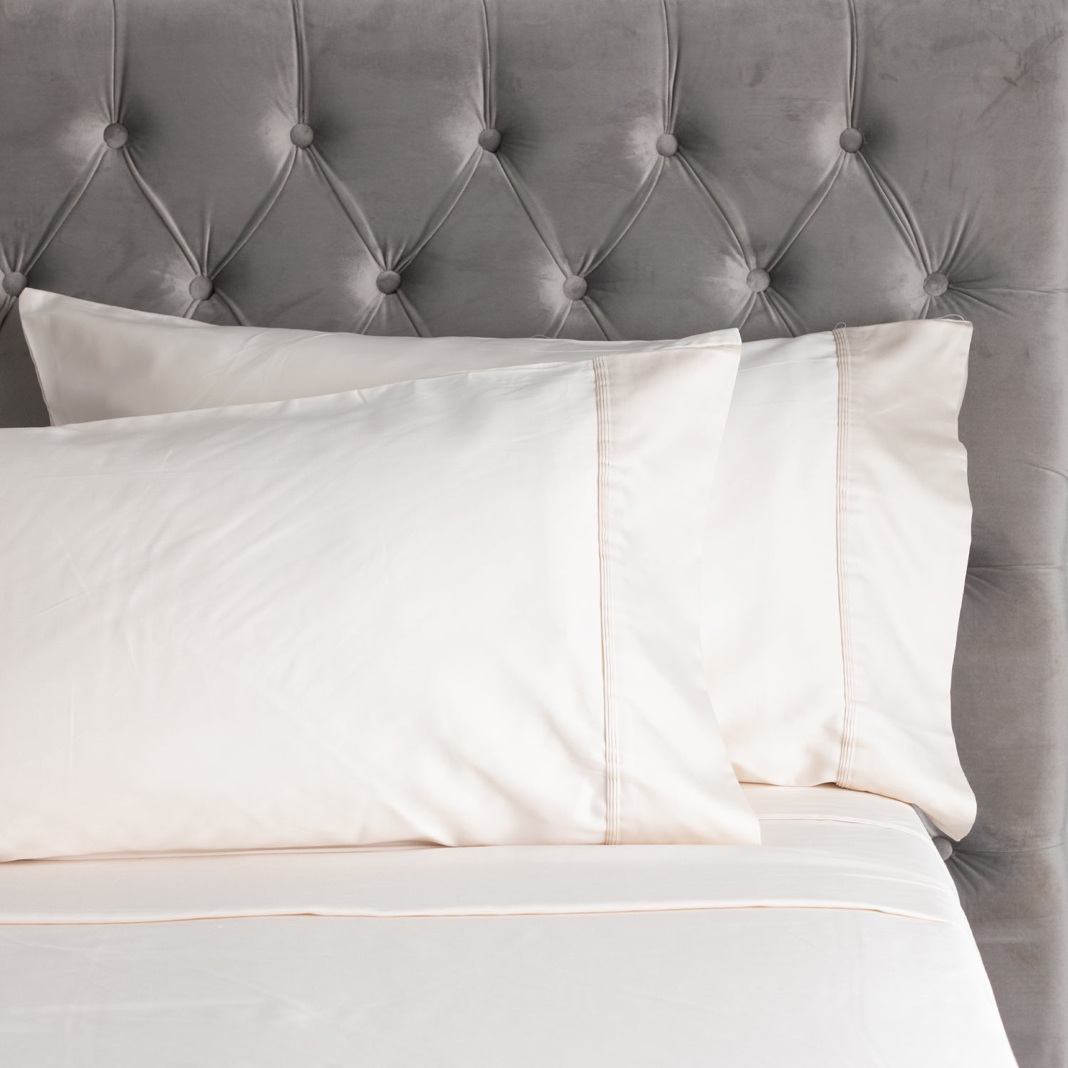 A close-up of smooth ivory sheets and matching pillowcases with elegant stitched cuff detailing, styled against a plush tufted velvet headboard.
