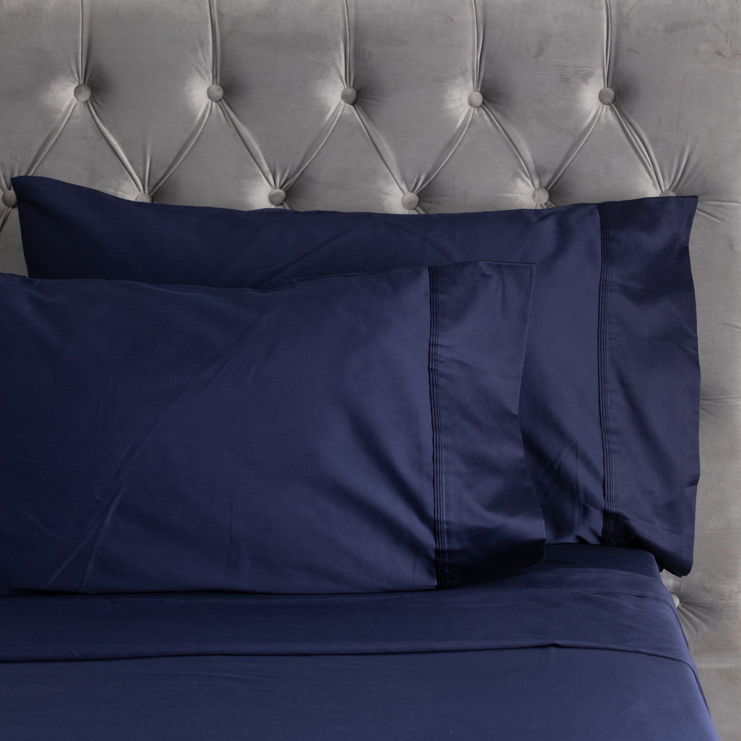 A close-up of smooth navy sheets and matching pillowcases with elegant stitched cuff detailing, styled against a plush tufted velvet headboard.