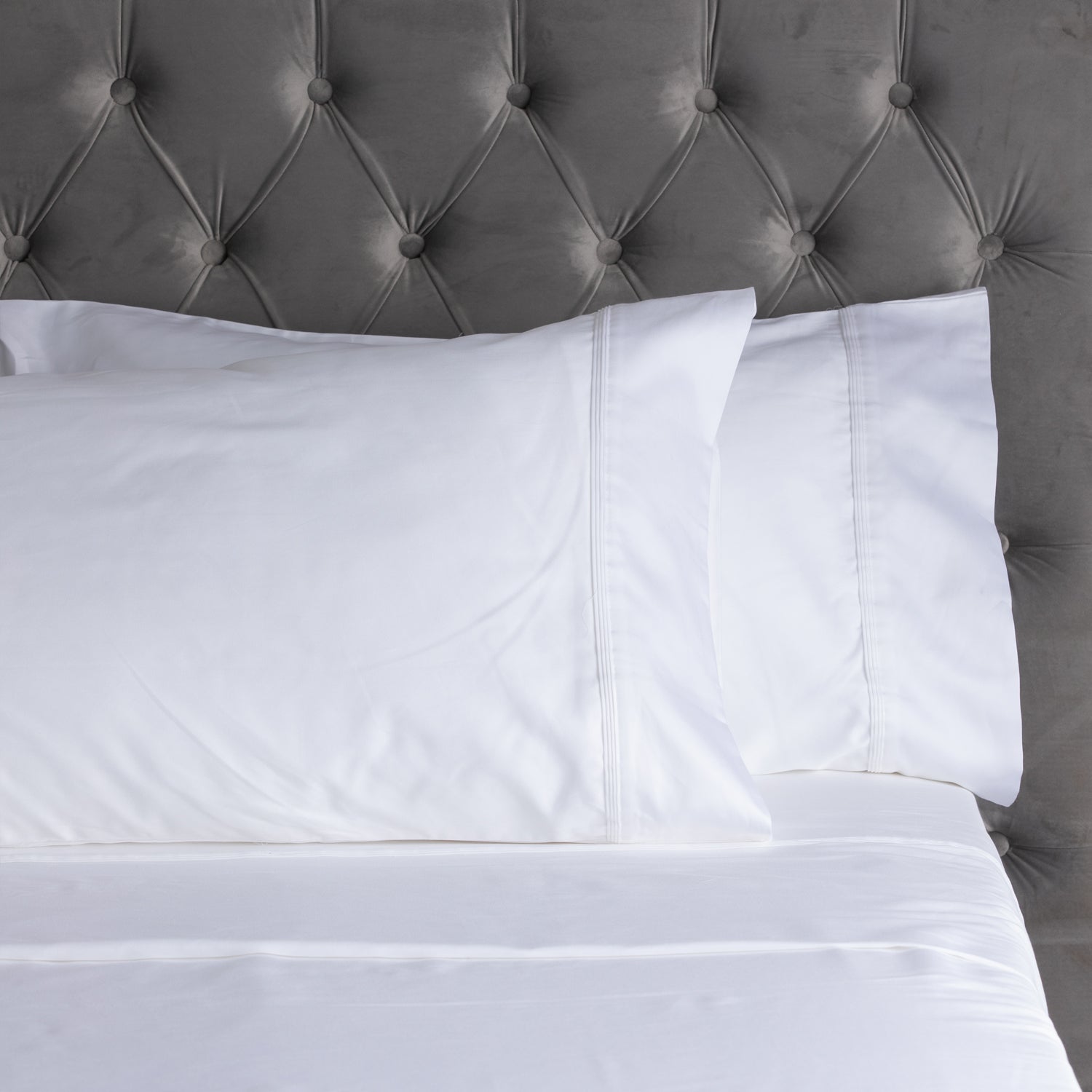 A close-up of smooth white sheets and matching pillowcases with elegant stitched cuff detailing, styled against a plush tufted velvet headboard.