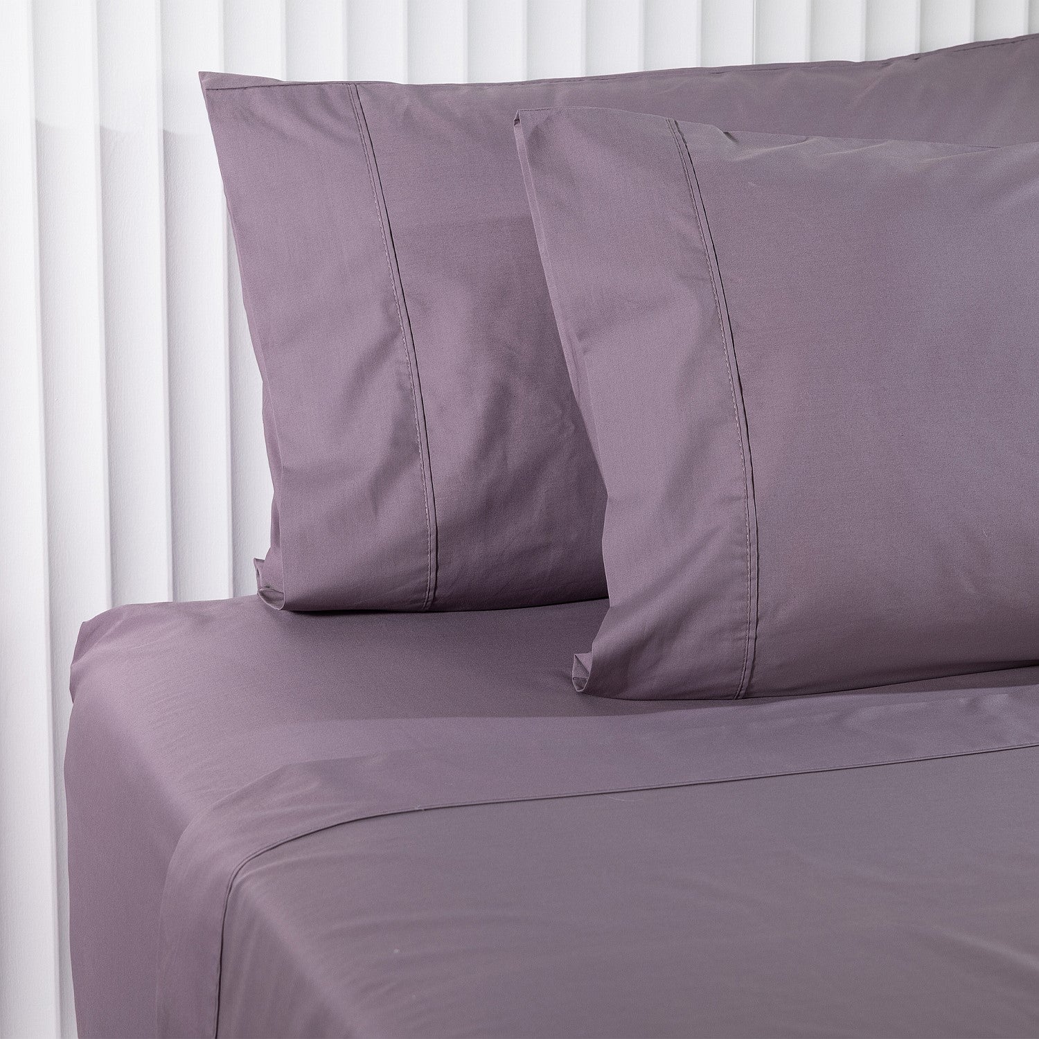 Purple bed sheets and pillowcases on a bed with white curtains in the background