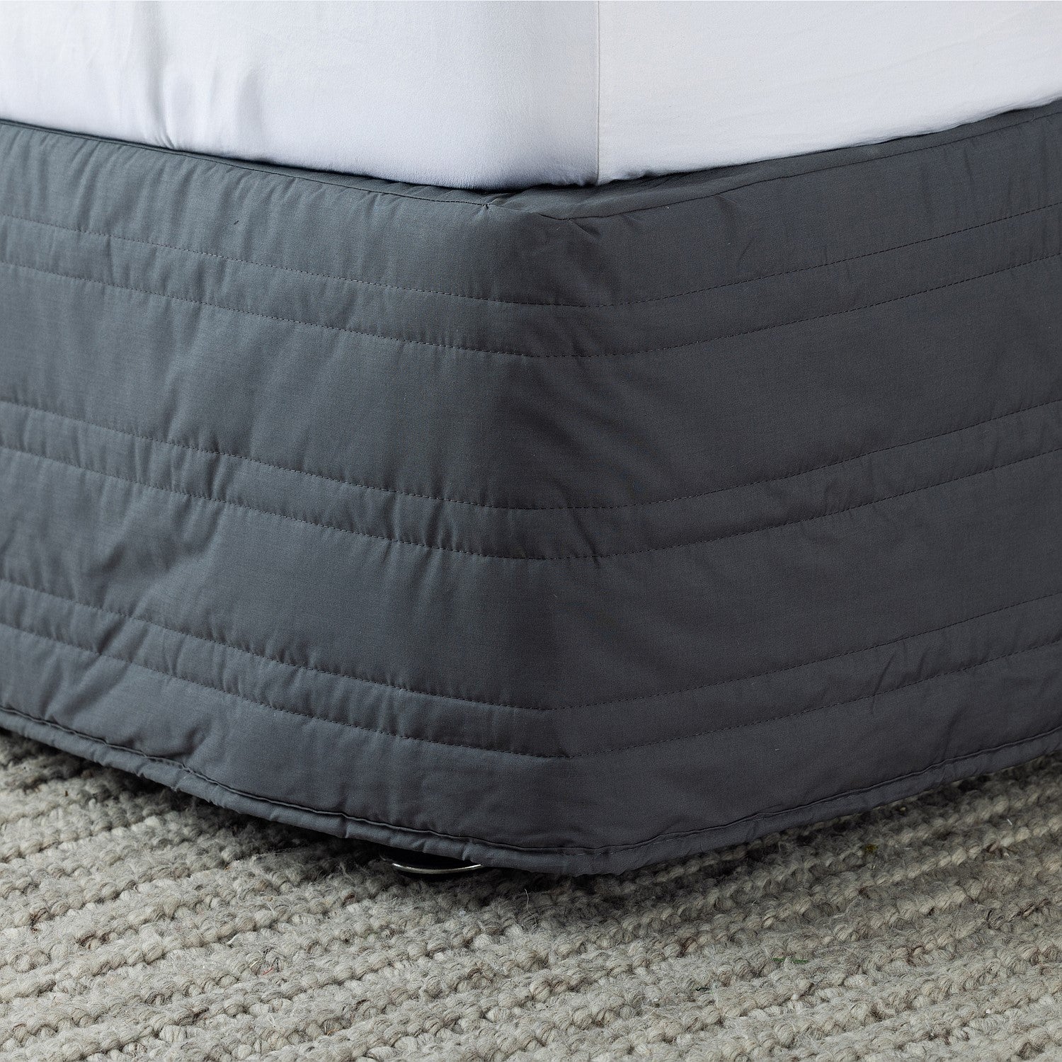 Gray quilted bed skirt on a bed with white bedding on a carpeted floor.