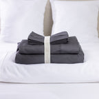 A premium charcoal sheet set is neatly folded and secured with a reusable branded storage band, beautifully displayed on a freshly made bed for a clean, luxurious look.