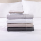 A neatly stacked collection of solid-colour sheet sets in white, silver, linen and charcoal showcases their smooth, luxurious finish against a softly blurred bedroom backdrop.