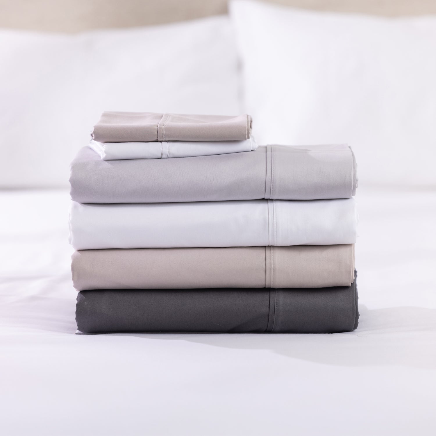 A neatly stacked collection of solid-colour sheet sets in white, silver, linen and charcoal showcases their smooth, luxurious finish against a softly blurred bedroom backdrop.