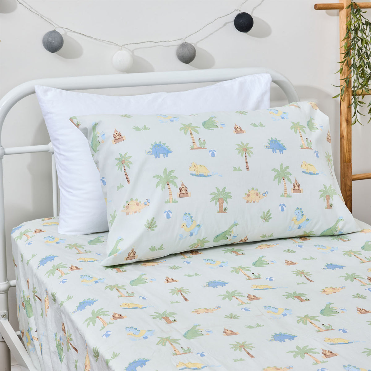 Close-up of the Dino Beach Party fitted sheet and pillowcase, showing colourful dinosaurs, palm trees, sandcastles and beach motifs on a pale blue background.