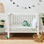 Dino Beach Party fitted sheet styled on a white toddler bed with a green dinosaur soft toy and white pillow.