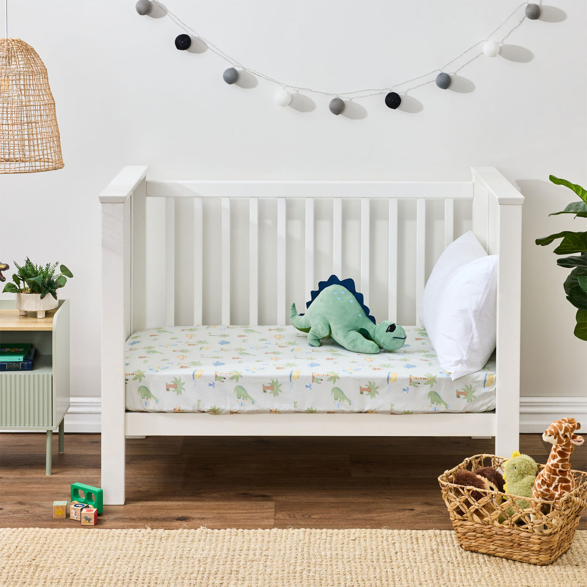 Dino Beach Party fitted sheet styled on a white toddler bed with a green dinosaur soft toy and white pillow.