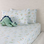 Dino Beach Party fitted sheet set styled on a bed with coordinating printed pillowcases and a green dinosaur soft toy.