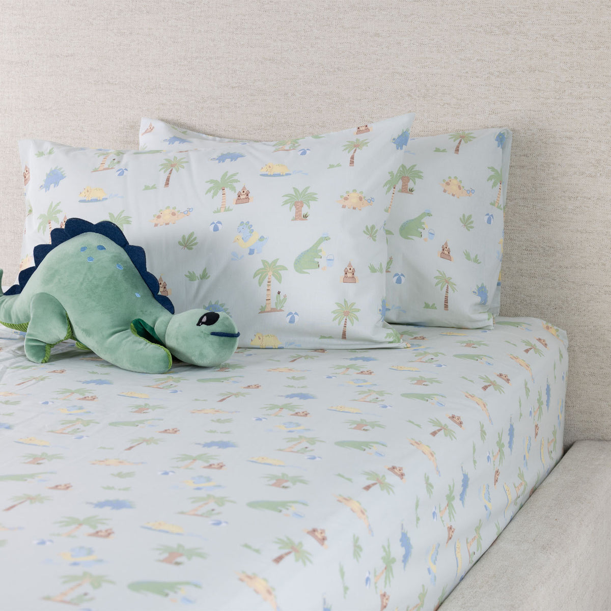 Dino Beach Party fitted sheet set styled on a bed with coordinating printed pillowcases and a green dinosaur soft toy.