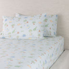 Dino Beach Party fitted sheet set displayed on a bed with coordinating pillowcases, showing the playful dinosaur and beach-themed print.