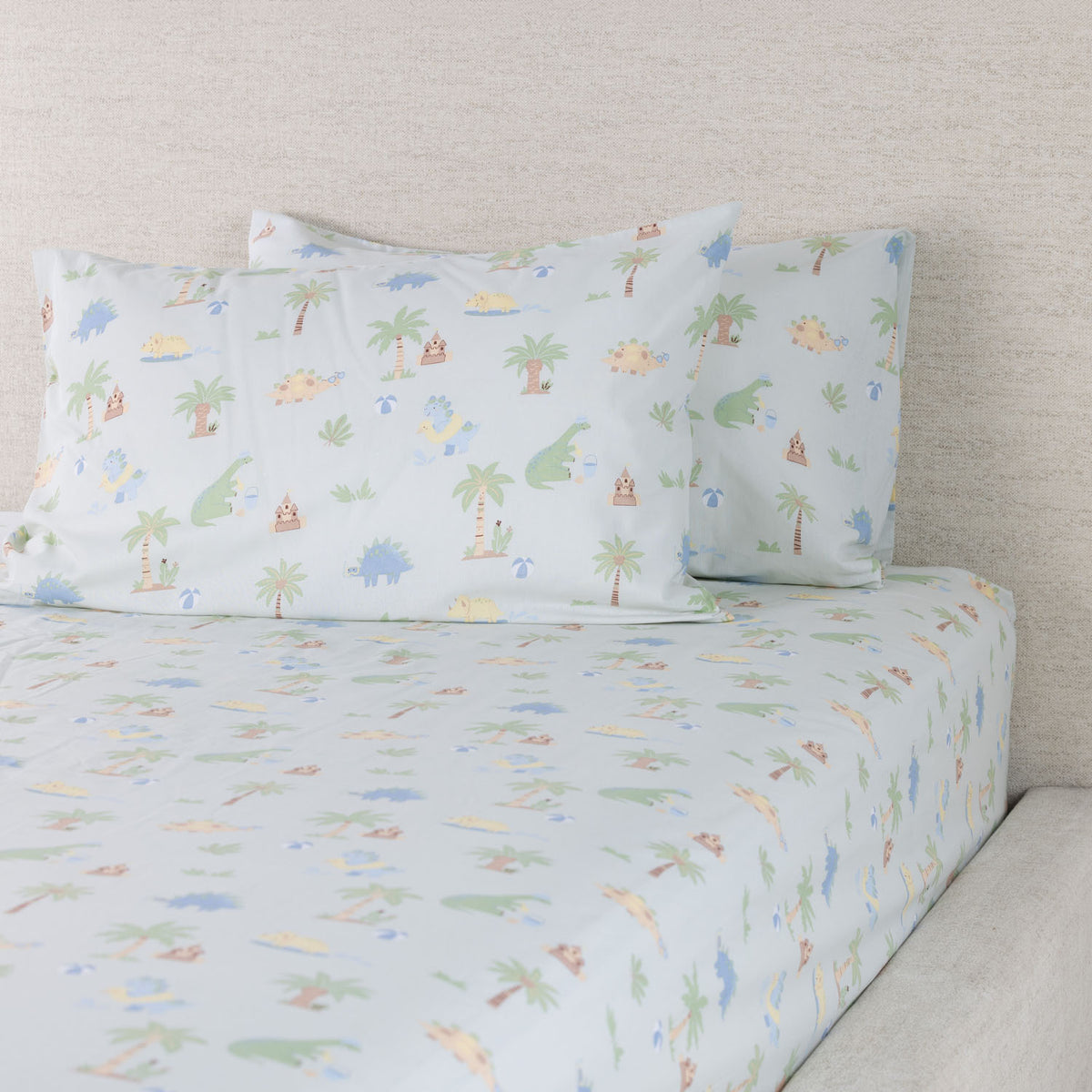 Dino Beach Party fitted sheet set displayed on a bed with coordinating pillowcases, showing the playful dinosaur and beach-themed print.