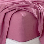 A berry linen fitted sheet is neatly wrapped around a mattress beneath softly draped linen bedding, showcasing its natural woven texture, snug fit and relaxed elegance.