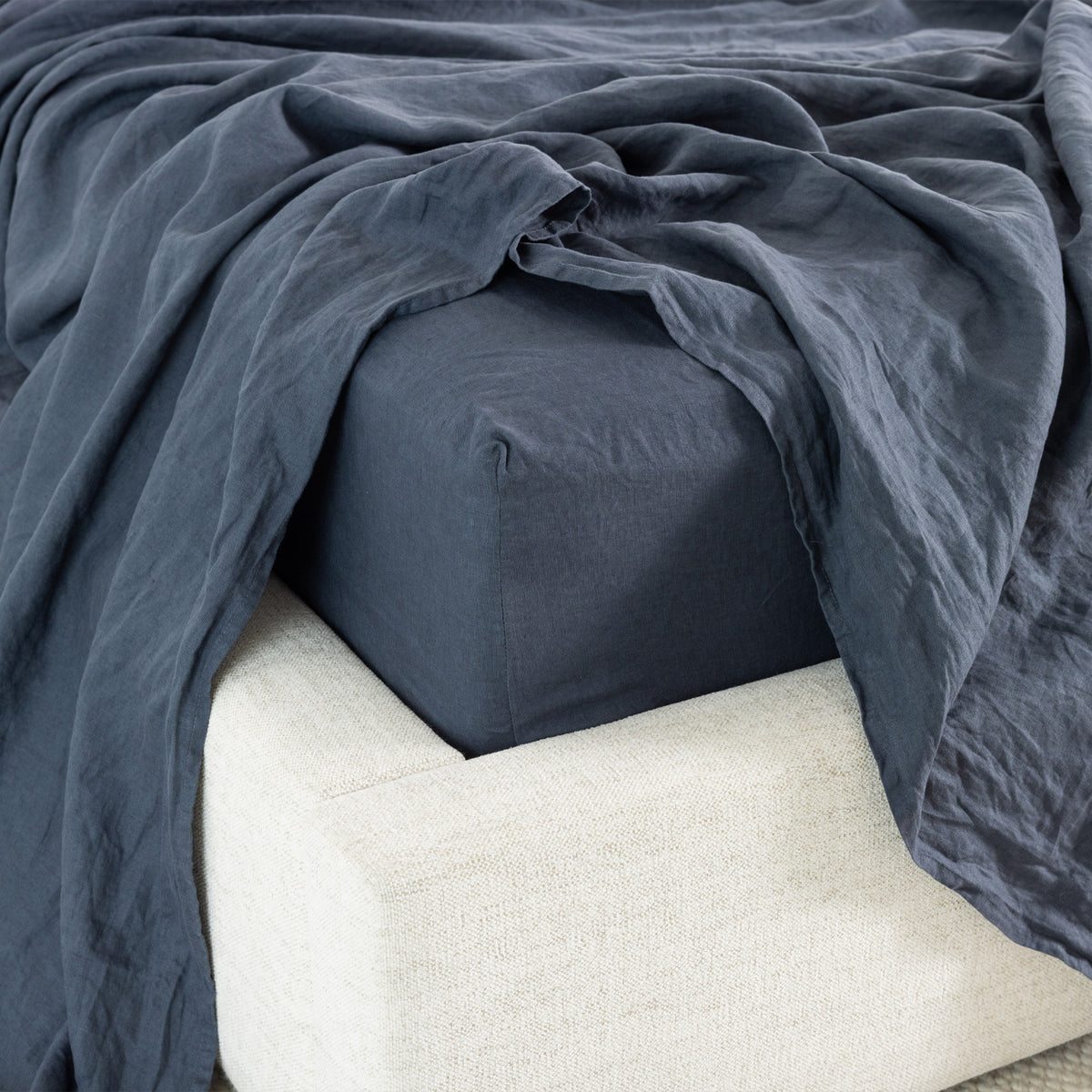 A midnight fitted sheet with a matching flat sheet on a bed, draped casually over the corner of the mattress over a light gray textured bed frame.