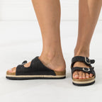 A pair of women's slide sandals with a black woven upper, double adjustable straps with silver buckles, and a cork-look midsole, shown on a model's feet against a white background.