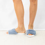A pair of fluffy chambray blue faux fur slippers is styled on foot, showcasing their plush texture, open-toe design and cosy everyday comfort.