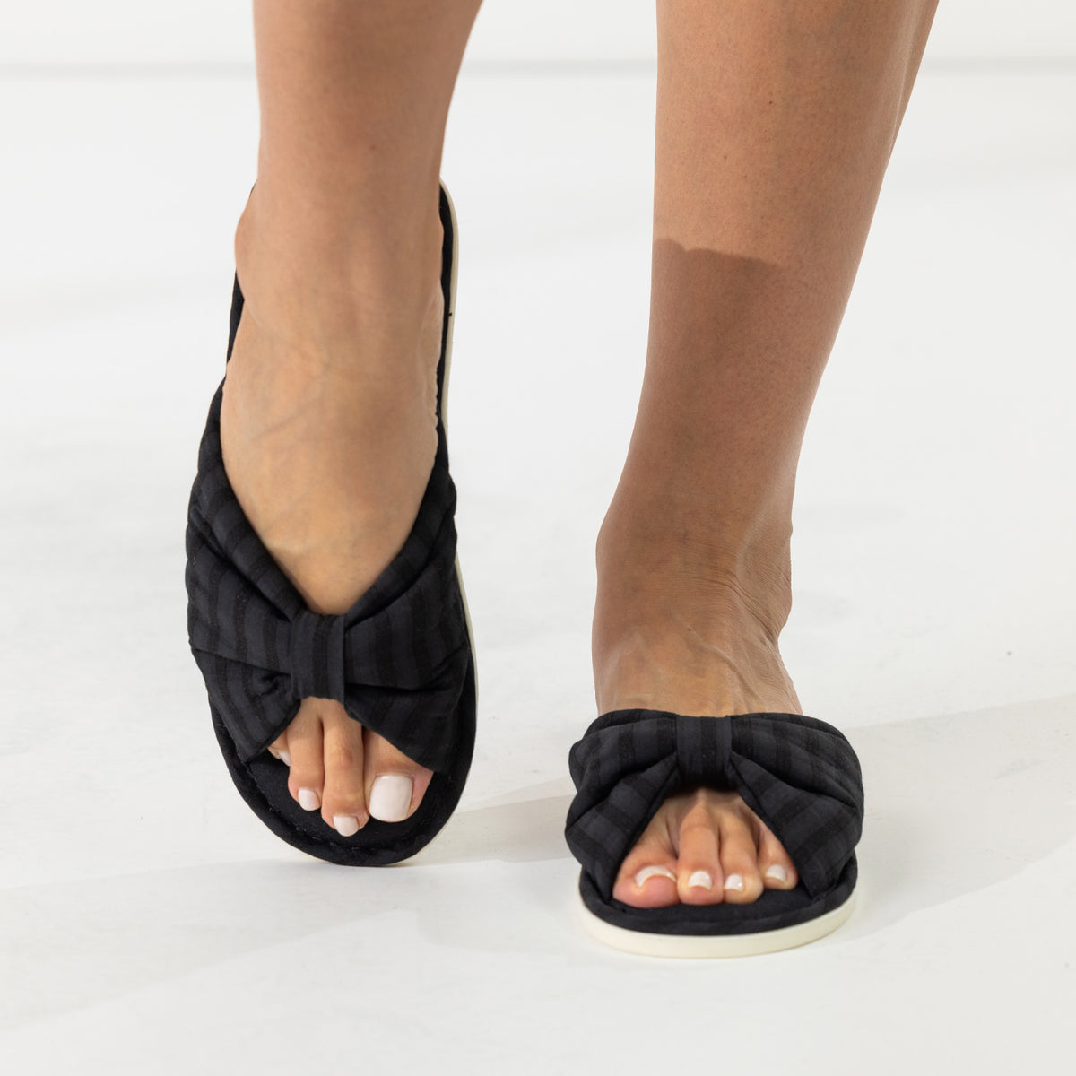 Black bow slippers shown while being worn, highlighting the soft padded straps and open-toe design.