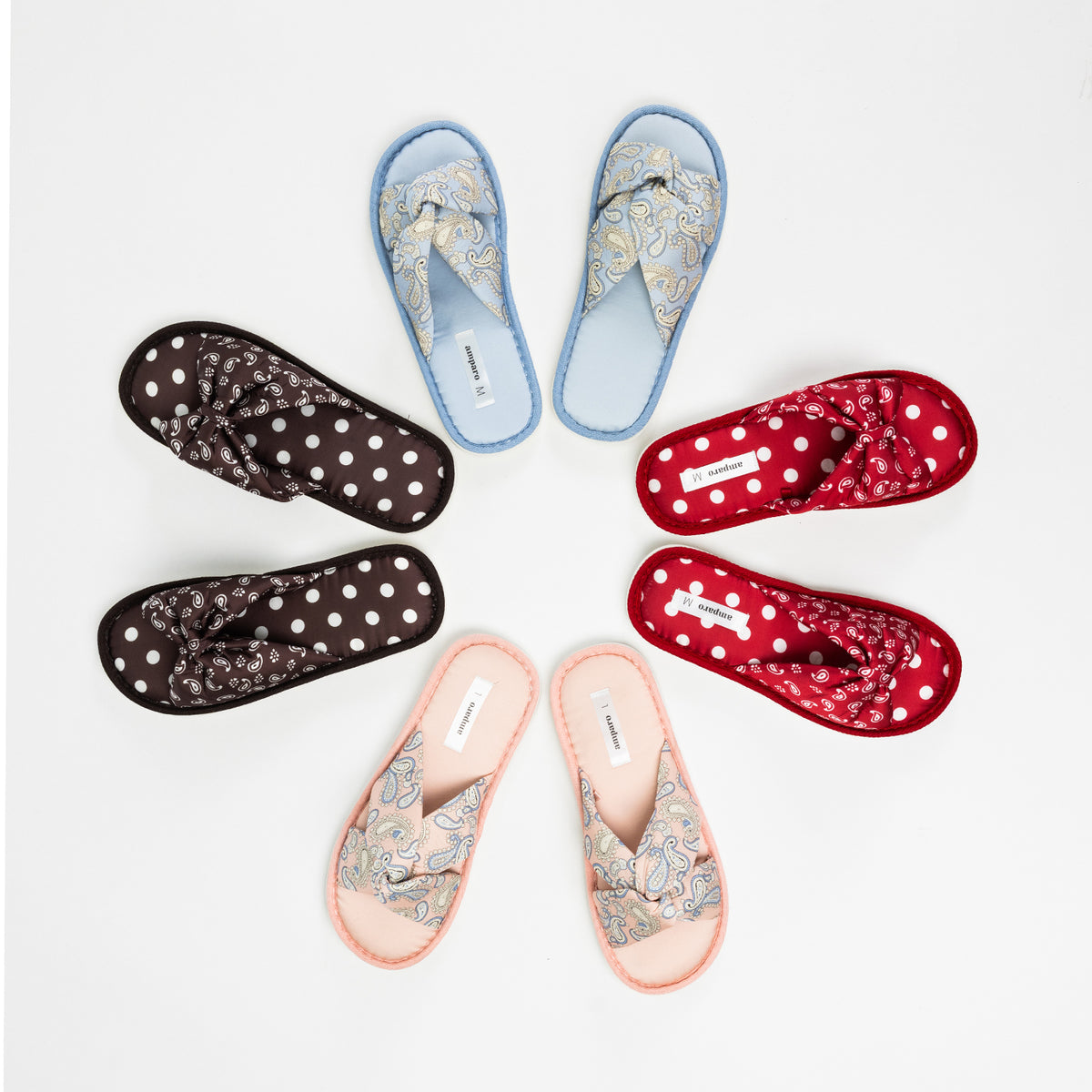 Overhead view of four pairs of Amparo open-toe slippers arranged in a circular display, featuring pink and denim blue paisley designs alongside raspberry red and chocolate brown polka dot designs, each with decorative crossover or bow-style straps on a white background.