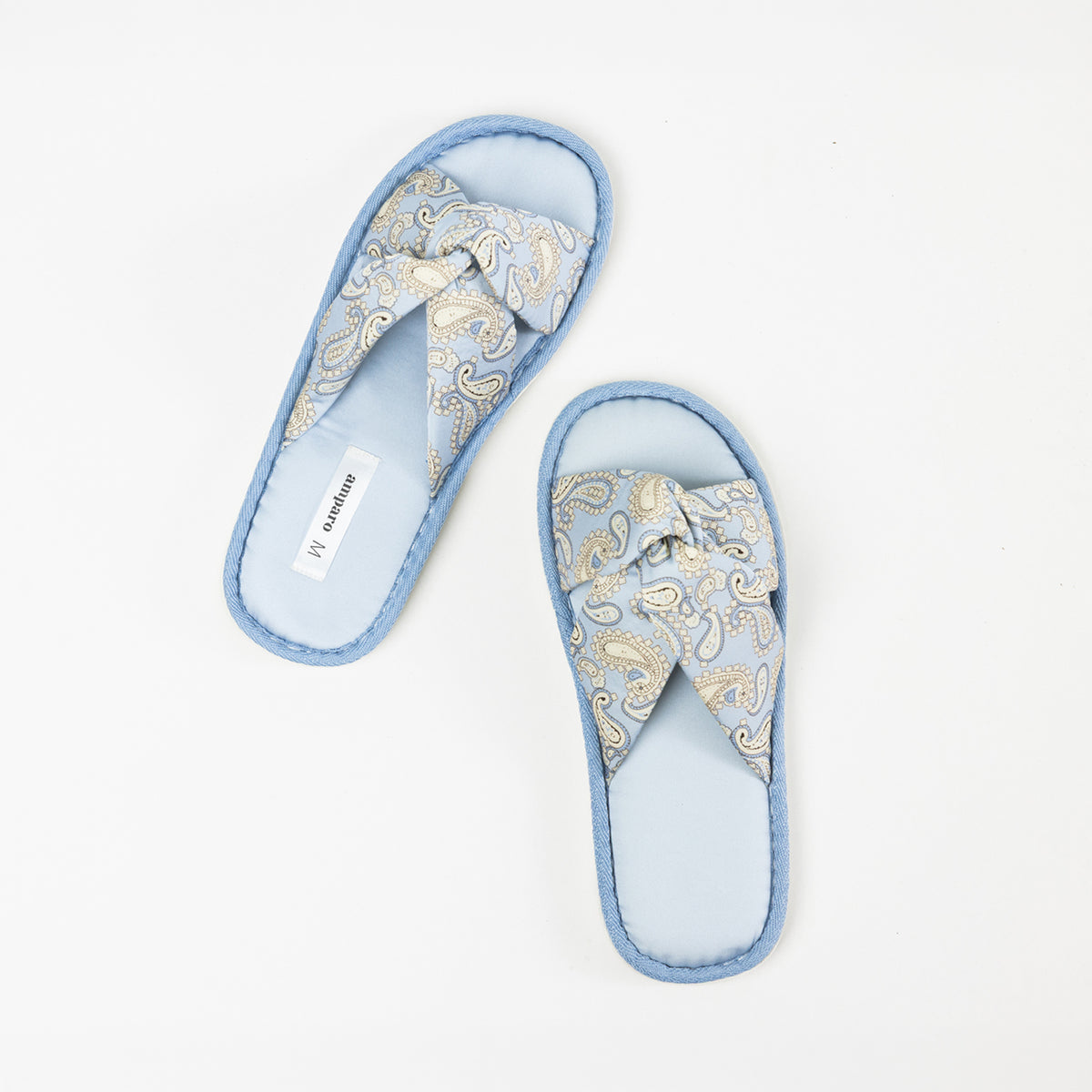 Top view of a pair of light blue open-toe slippers featuring cushioned footbeds, crossover straps with a cream and blue paisley bandana print, and coordinating denim blue trim on a white background.