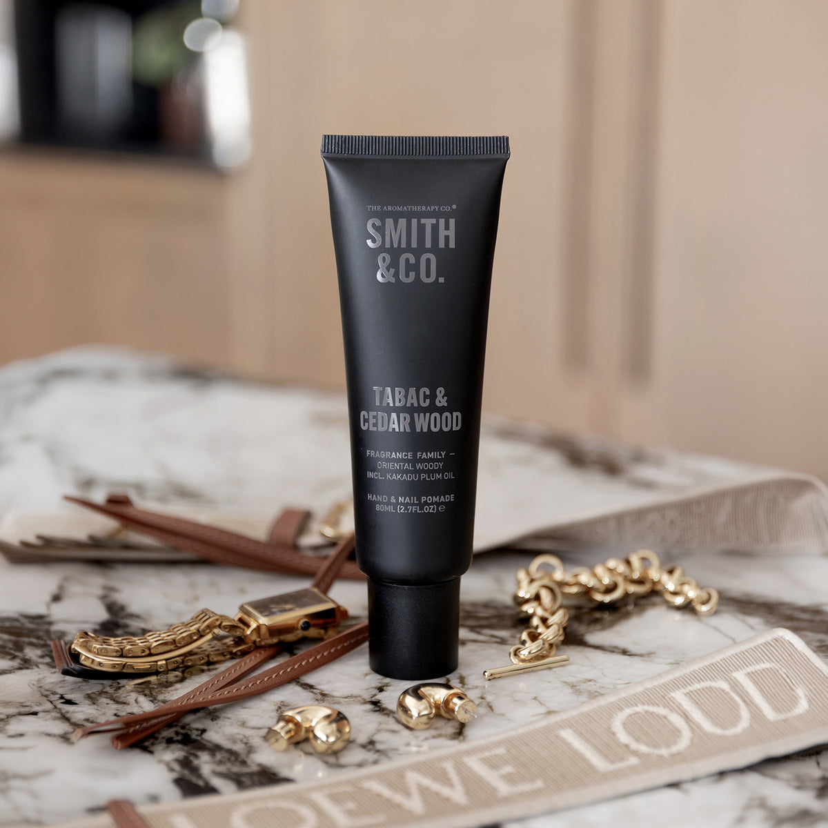 Smith & Co. Tabac & Cedarwood Hand & Nail Pomade 80ml standing on a marble surface surrounded by gold jewellery and tan leather accessories.