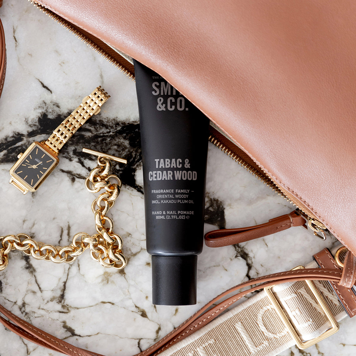Smith & Co. Tabac & Cedarwood Hand & Nail Pomade 80ml nestled beside a tan leather handbag, gold jewellery and a watch on a marble surface.