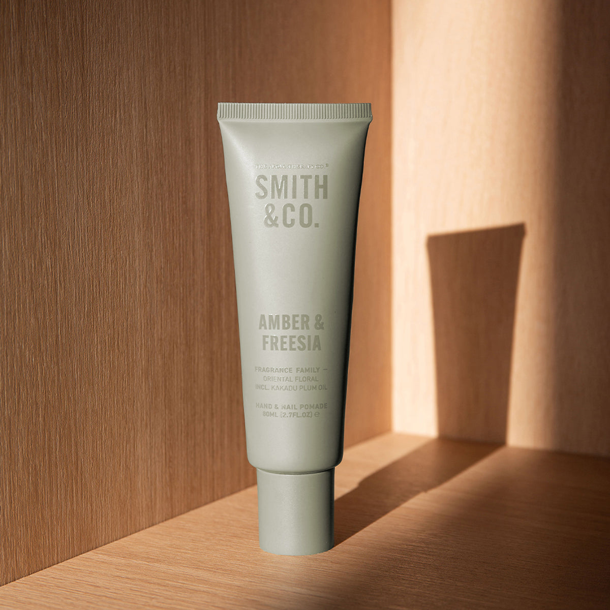 Smith & Co. Amber & Freesia Hand & Nail Pomade 80ml displayed on a wooden shelf in warm sunlight with soft shadows.