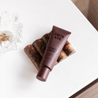 Smith & Co. Black Oud & Saffron Hand & Nail Pomade 80ml in a burgundy tube resting on a decorative brown stone tray beside gold jewellery.