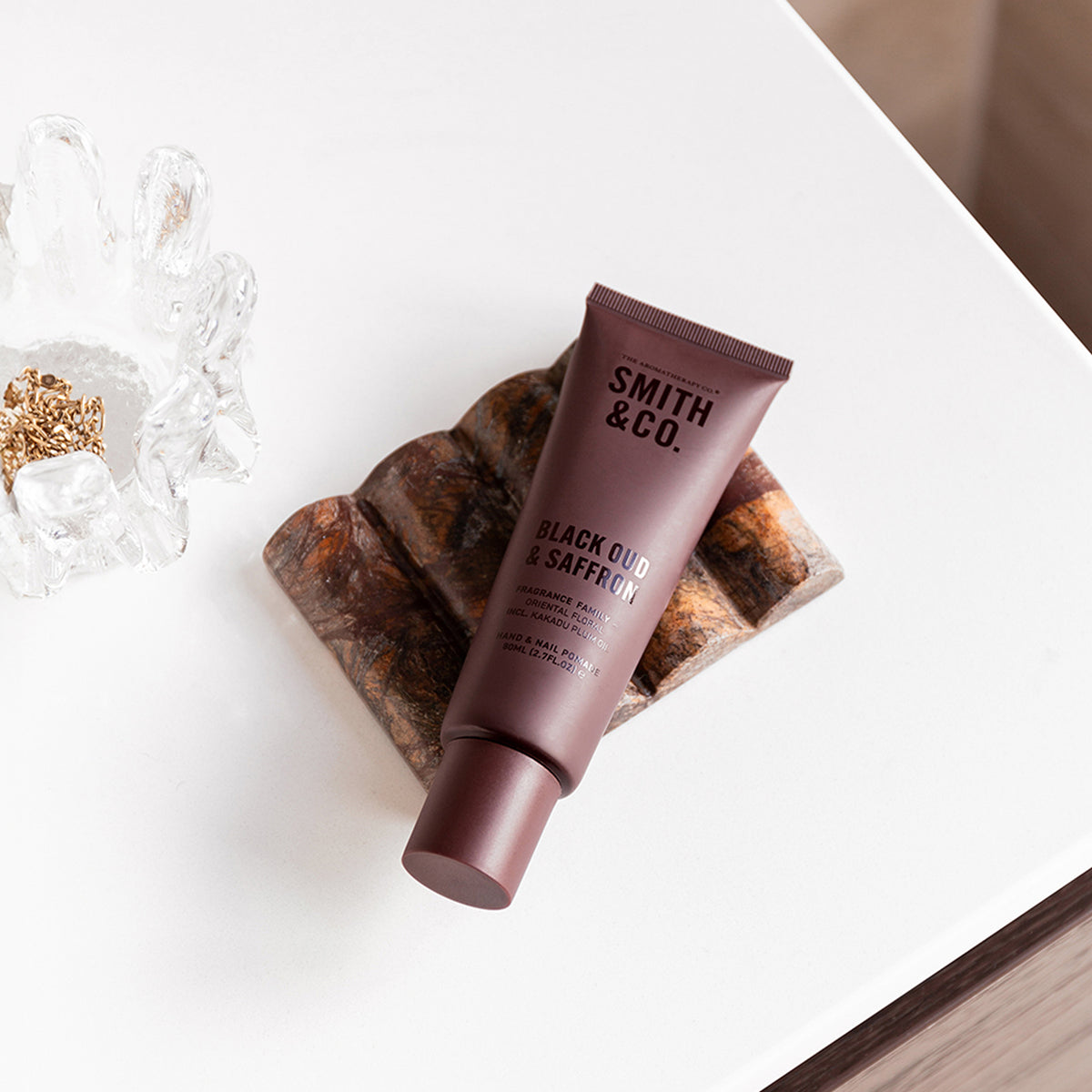 Smith & Co. Black Oud & Saffron Hand & Nail Pomade 80ml in a burgundy tube resting on a decorative brown stone tray beside gold jewellery.