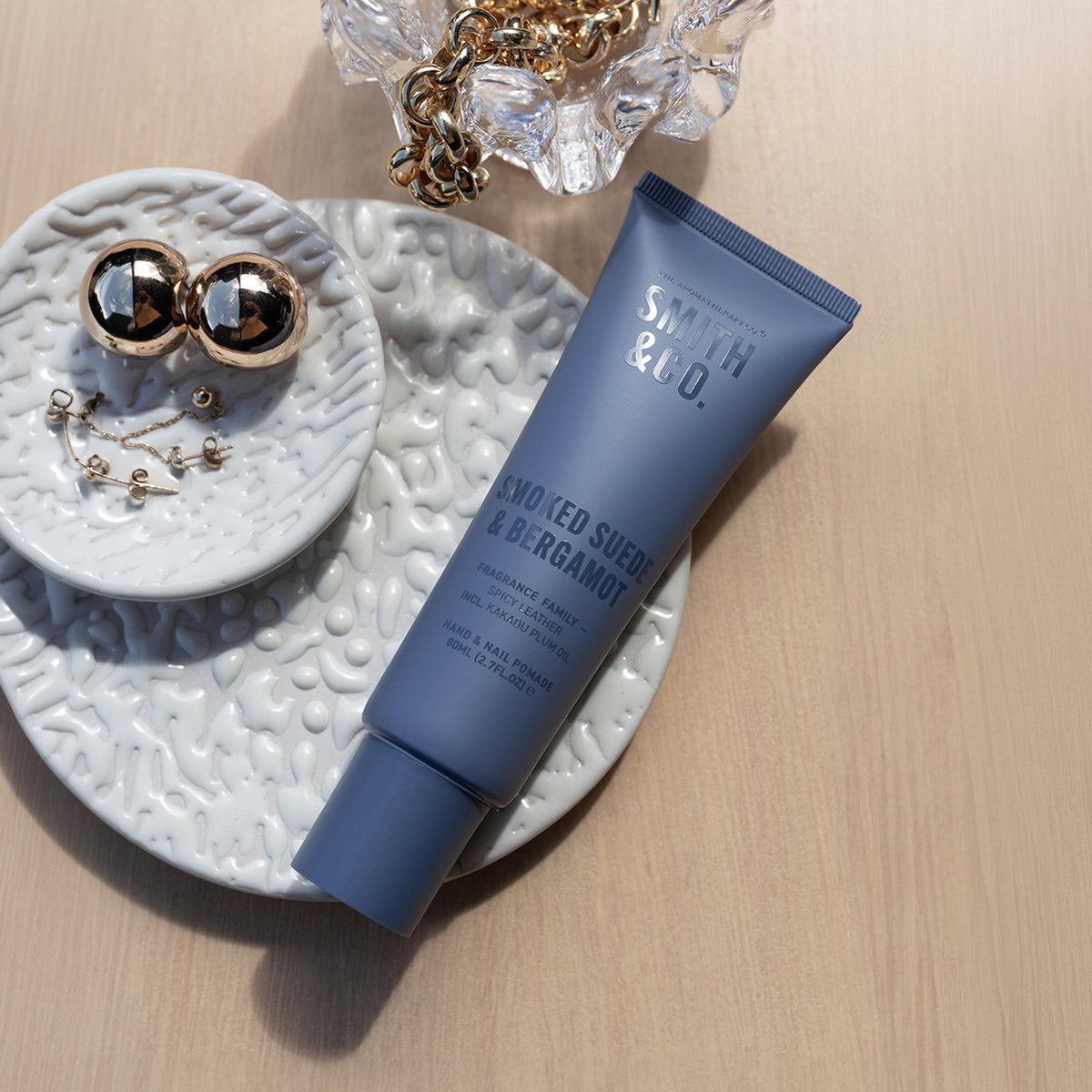 Smith & Co. Smoked Suede & Bergamot Hand & Nail Pomade 80ml displayed on a white textured tray alongside gold jewellery and decorative accessories.