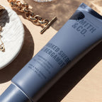 Smith & Co. Smoked Suede & Bergamot Hand & Nail Pomade 80ml in a dusty blue tube resting on a beige surface beside gold jewellery and a white decorative dish.