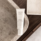 A white squeeze tube of Smith & Co. Tonka & White Musk fragrance family hand and heel powder stands on a curved stone surface with a bathroom-like setting in the background.