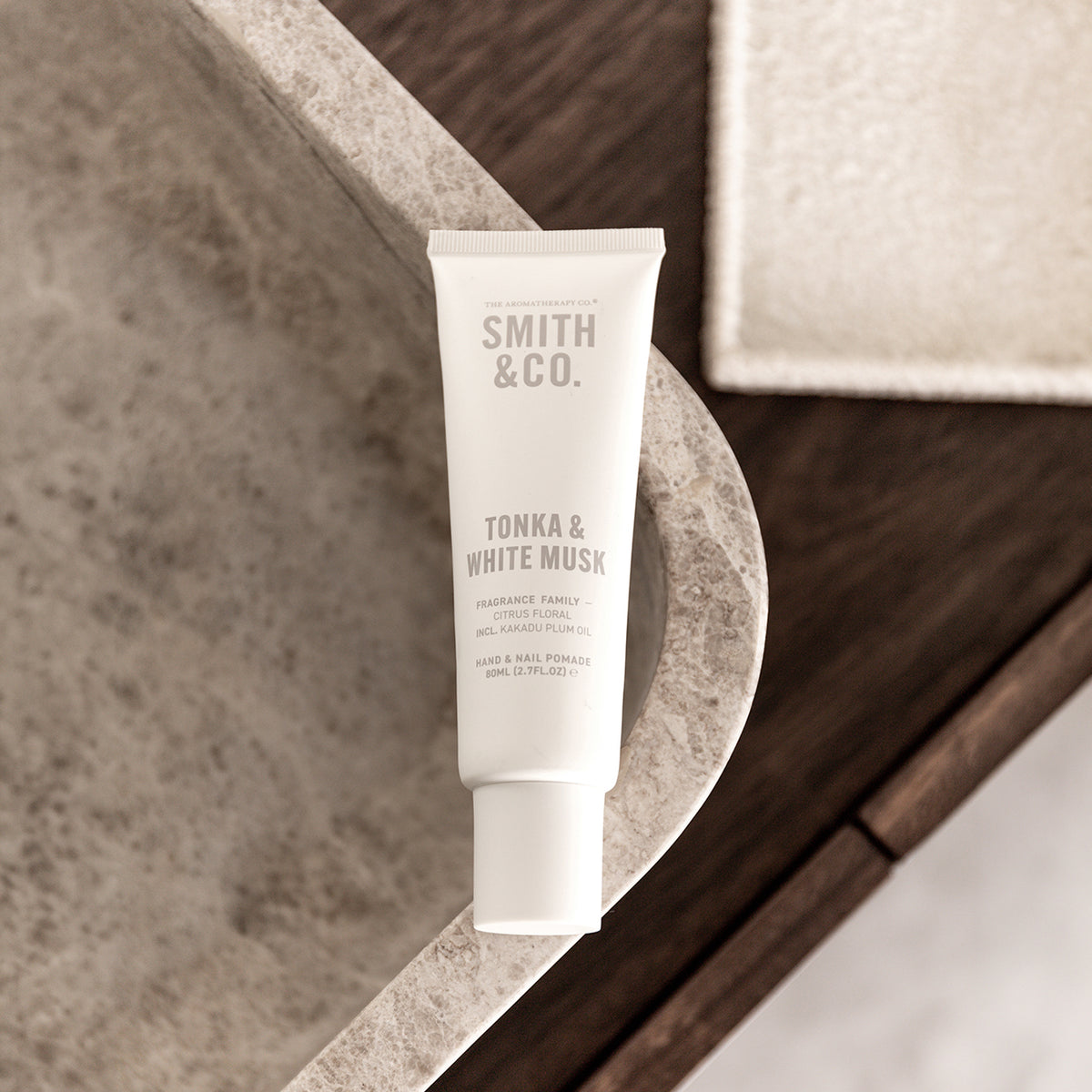 A white squeeze tube of Smith & Co. Tonka & White Musk fragrance family hand and heel powder stands on a curved stone surface with a bathroom-like setting in the background.