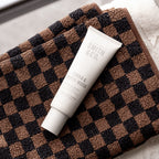 A white tube of Smith & Co. Tonka & White Musk facial cleanser resting diagonally on a brown and black checkered plush towel.