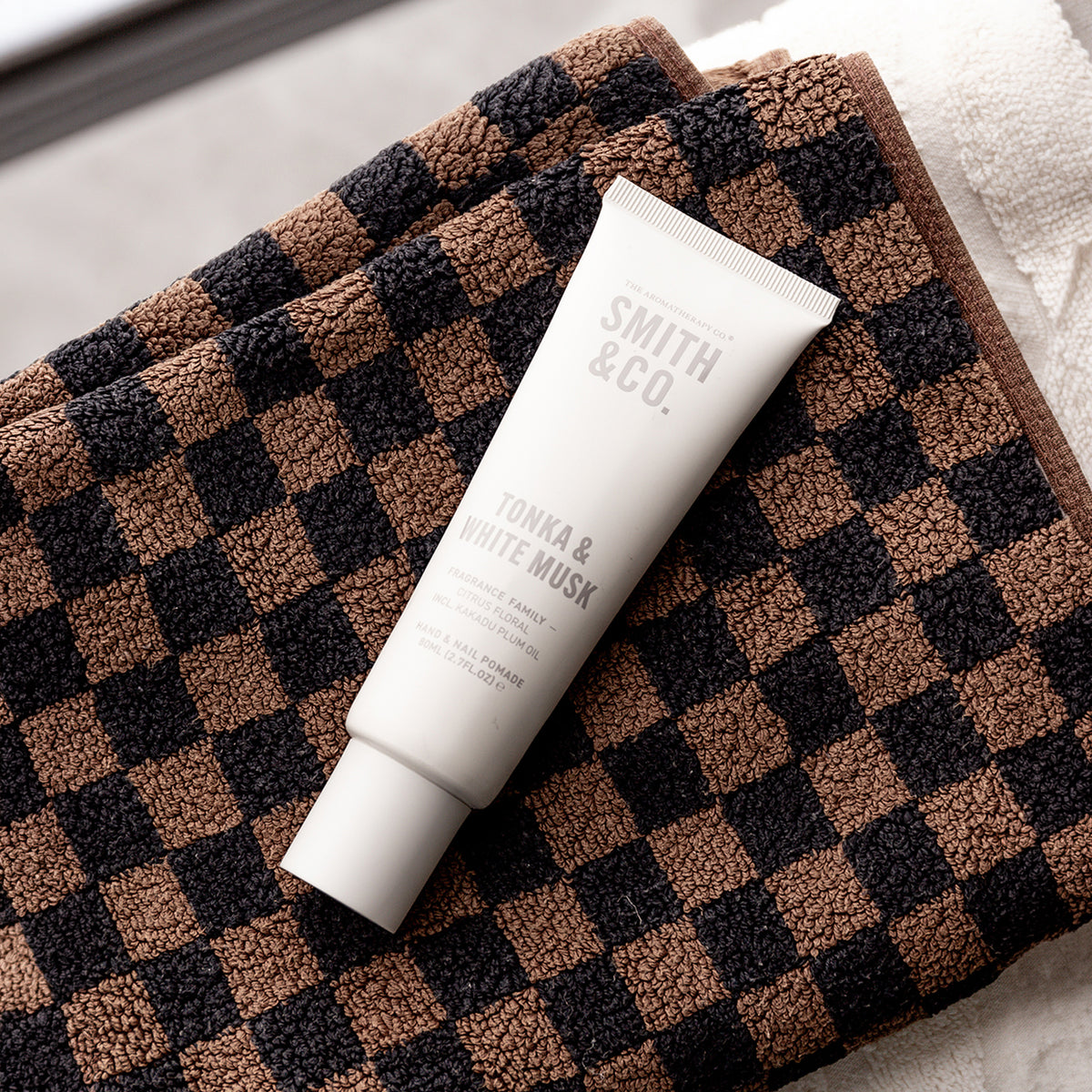 A white tube of Smith & Co. Tonka & White Musk facial cleanser resting diagonally on a brown and black checkered plush towel.