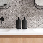 Two matte black Smith & Co. pump bottles positioned between bathroom sinks on a white vanity with grey terrazzo tiles and timber cabinetry.