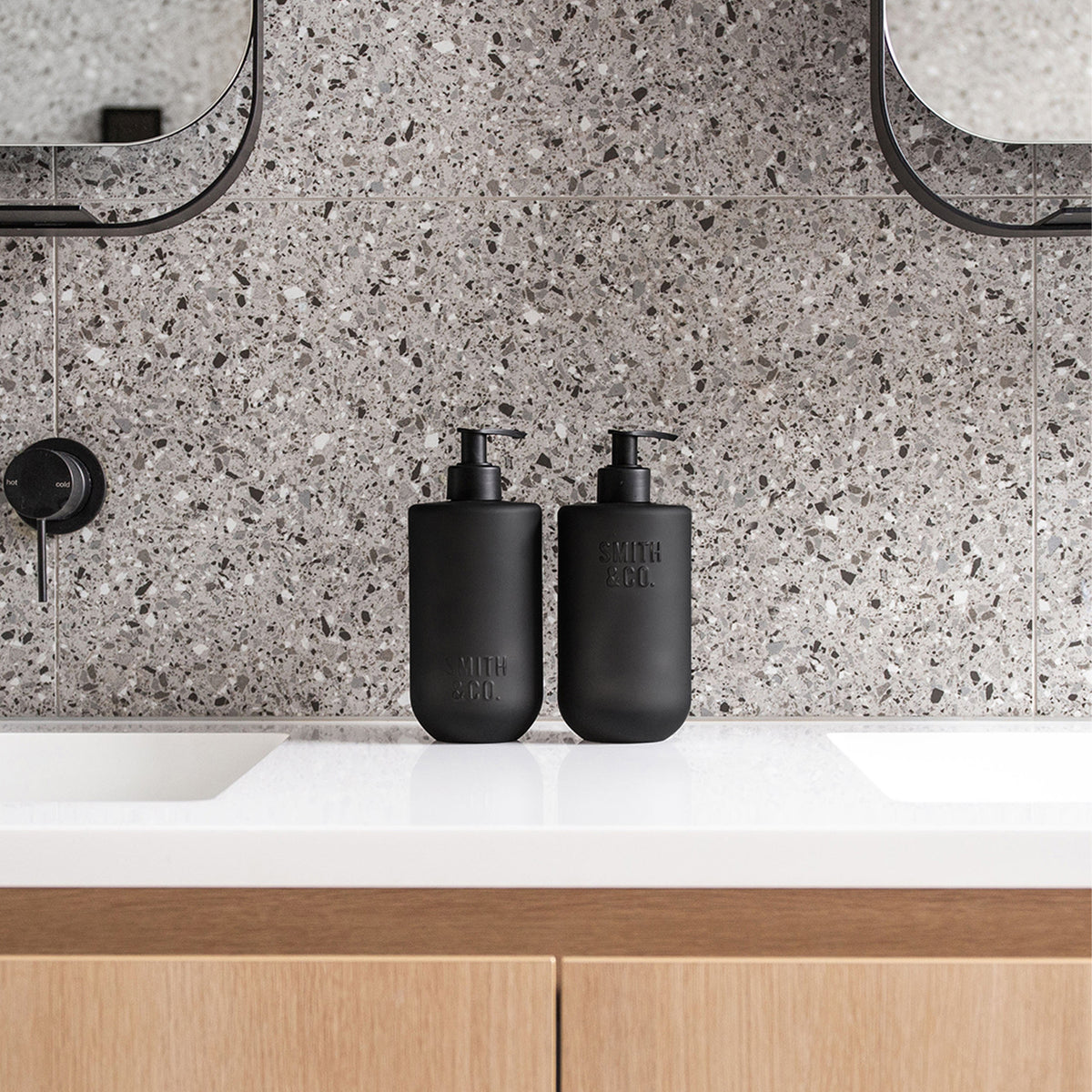 Two matte black Smith & Co. pump bottles positioned between bathroom sinks on a white vanity with grey terrazzo tiles and timber cabinetry.