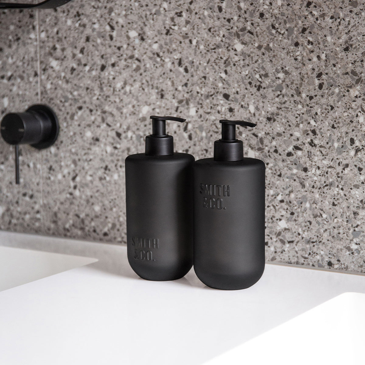 Two matte black Smith & Co. pump bottles displayed on a white bathroom countertop against a grey terrazzo tiled wall.